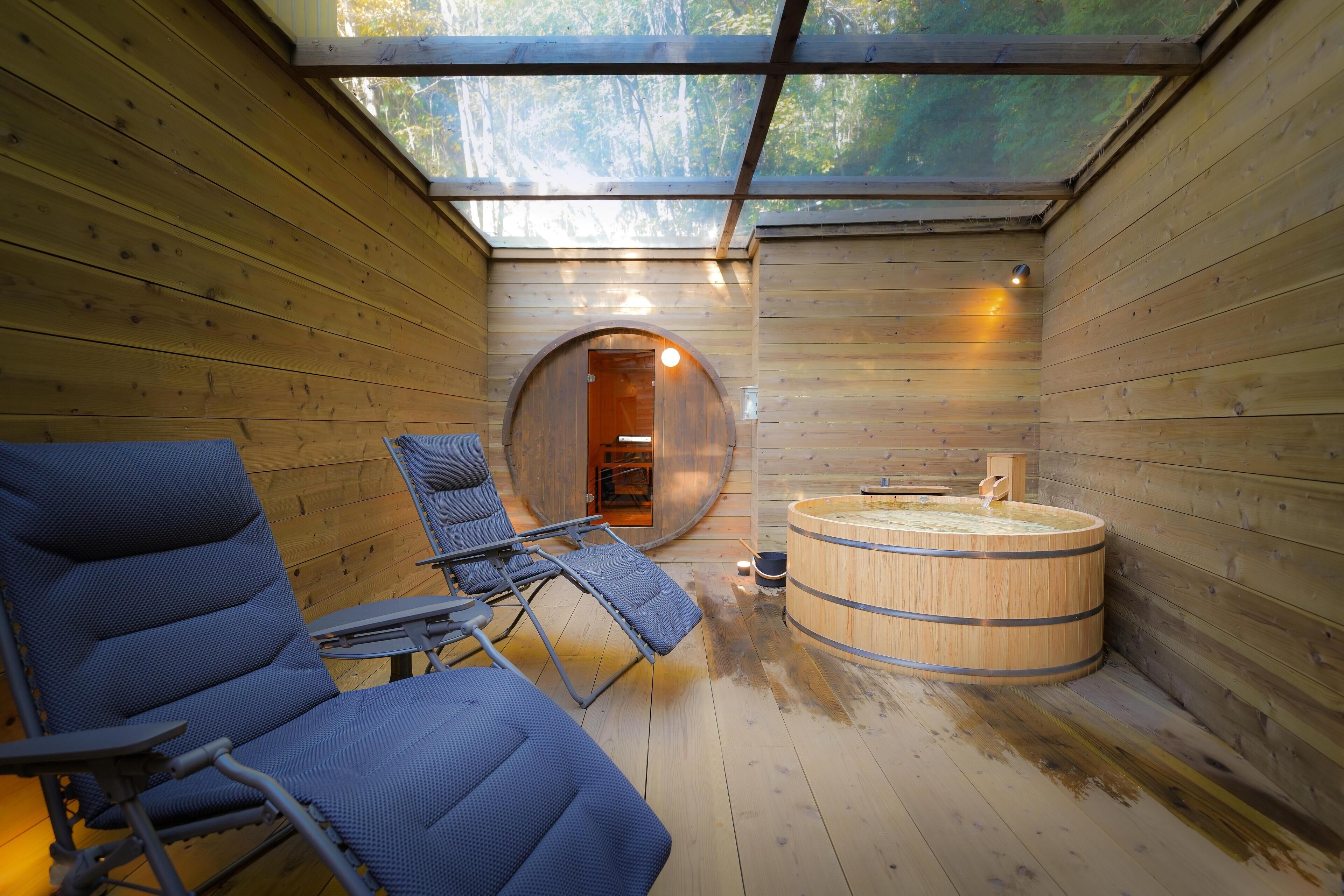 A wooden cabin with a glass ceiling, a round wooden bathtub, and blue cushioned chairs.