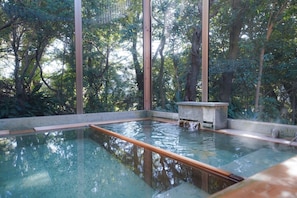 Una piscina cristalina con bordes de madera, rodeada de árboles altos y una estructura de piedra, probablemente un área de aguas termales o baños.