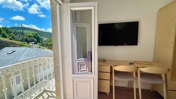 A balcony with a white railing, a wooden desk, a chair, a television mounted on the wall, and a view of a green landscape with houses.