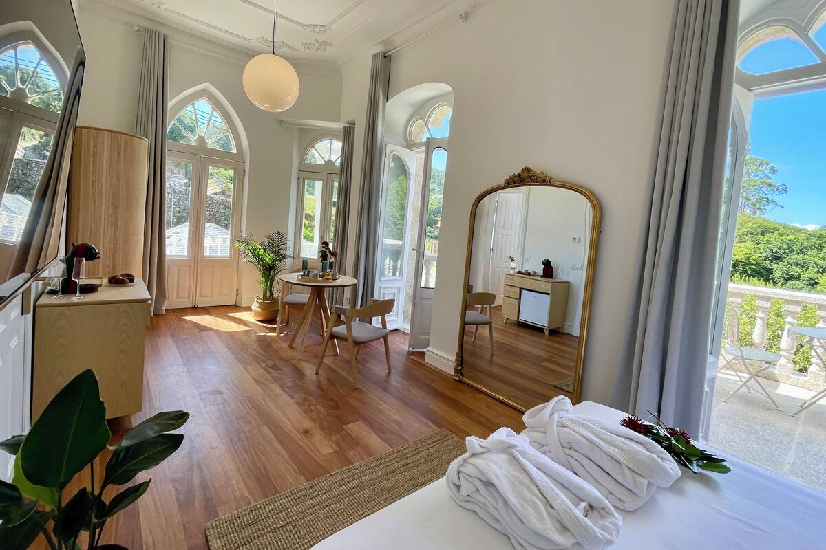 Ein geräumiges Zimmer mit einem großen Bett, einem Schreibtisch, einem Stuhl und einem Spiegel. Große Fenster und ein Balkon sind zu sehen.
