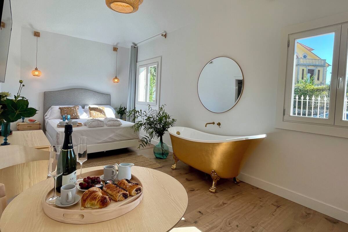 Ein modernes Schlafzimmer mit einem Bett, einer goldenen Badewanne, einem runden Spiegel und einem Frühstücksbrett mit Croissants und Weingläsern.
