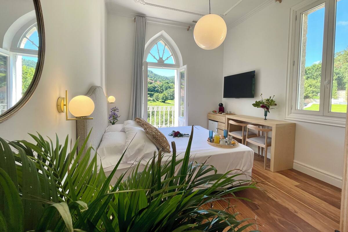 Ein Schlafzimmer mit einem Bett, einem Nachttisch mit einem Frühstücksbrett, einem Fernseher und einem großen Fenster mit Blick auf Grünflächen.