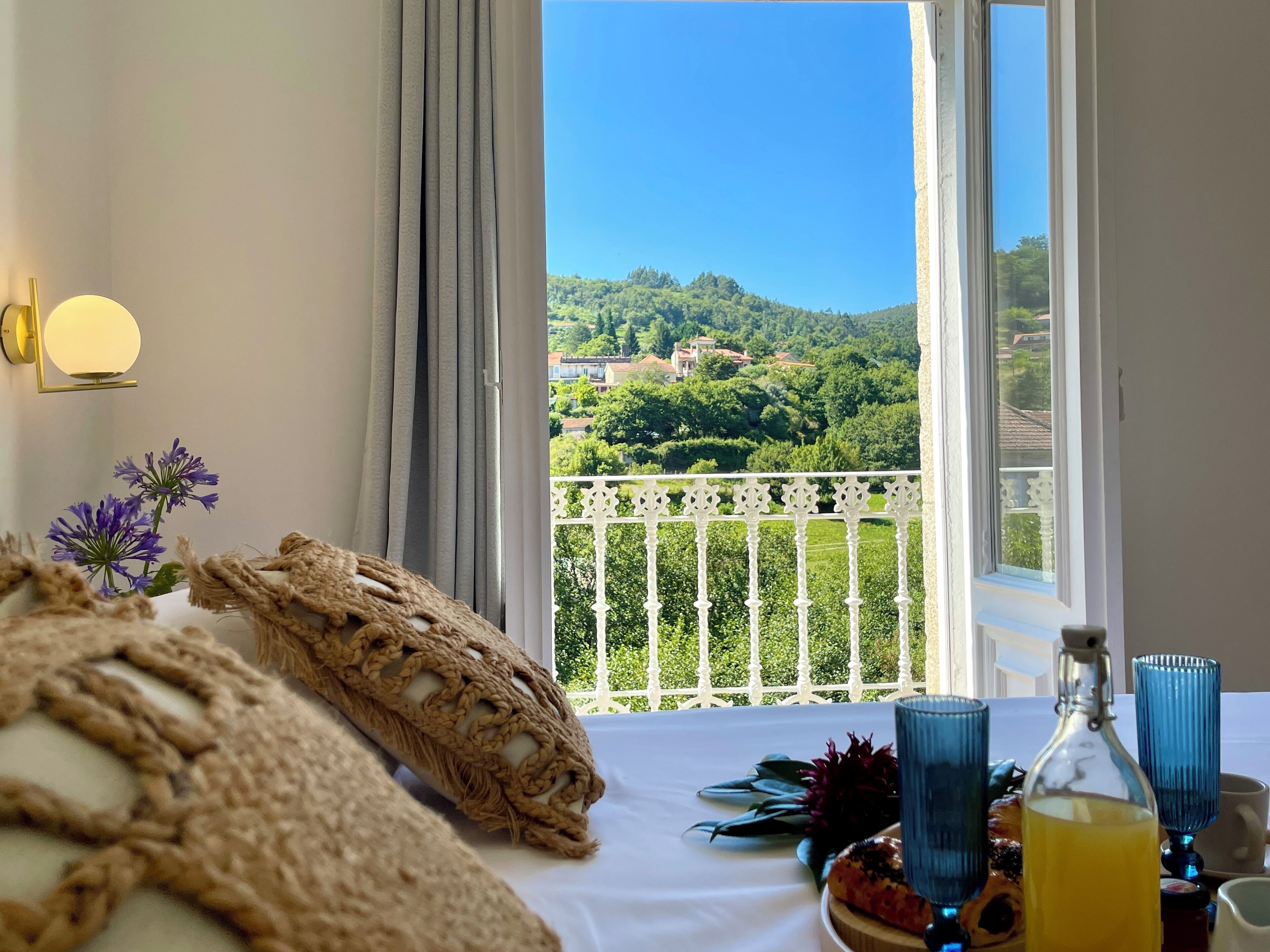 A room with a view of a green landscape, a white balcony railing, and a table with a bottle and glasses.