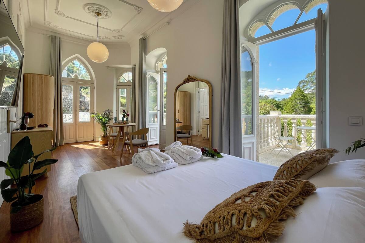 Ein geräumiges Zimmer mit einem großen Bett, einem Holztisch, einem Stuhl und einem Balkon mit Geländer.