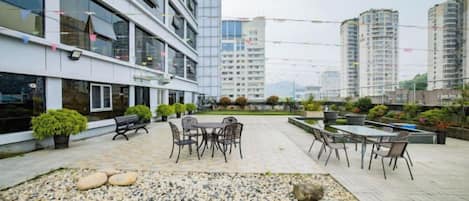 Una terraza al aire libre con mobiliario, un pequeño jardín y edificios modernos al fondo.