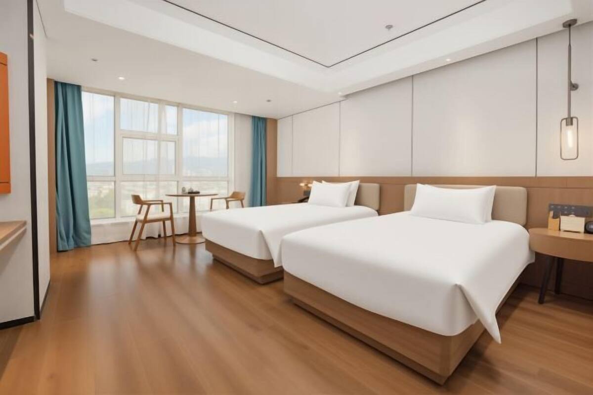 Ein Hotelzimmer mit zwei Betten, einem kleinen Tisch, einem Stuhl und einem großen Fenster mit Vorhängen.
