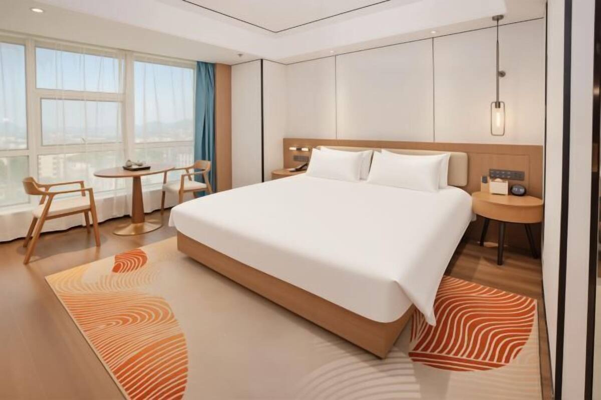 Ein modernes Hotelzimmer mit einem großen Bett, einem kleinen Tisch mit Stühlen, einem Nachttisch und einem großen Fenster mit Vorhängen.