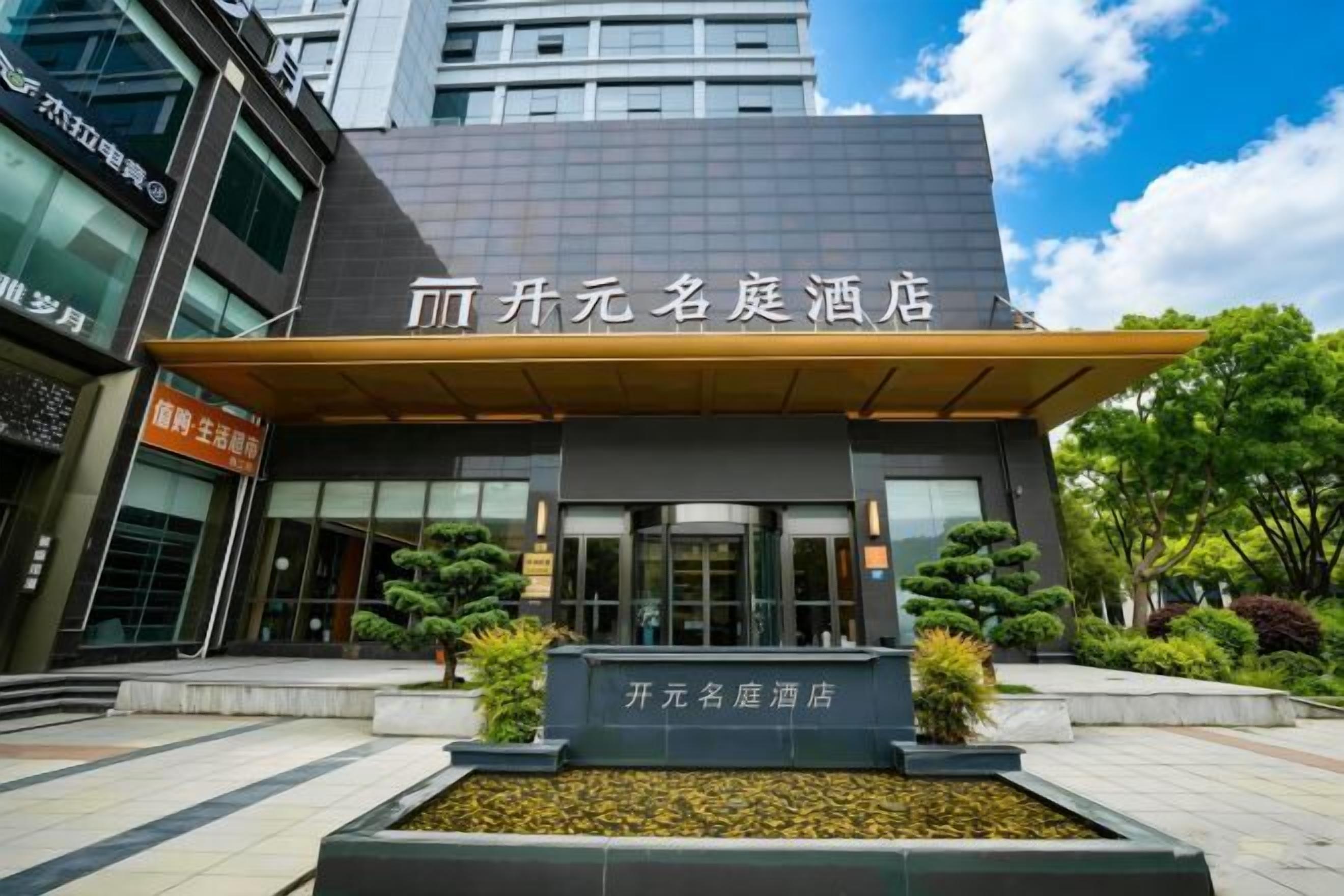 A hotel entrance with a modern facade, a signboard, and a landscaped area with plants.