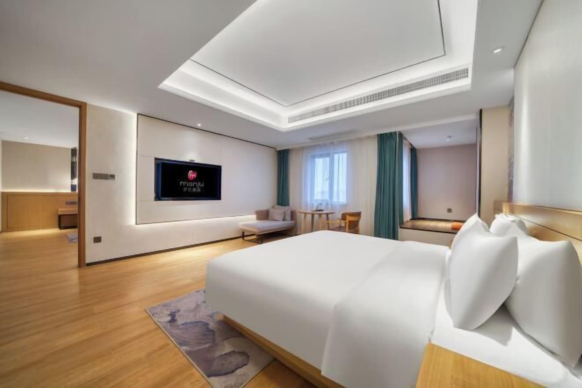 A modern hotel room with a large bed, a flat-screen TV mounted on the wall, a small seating area with a table and chairs, and a large mirror.