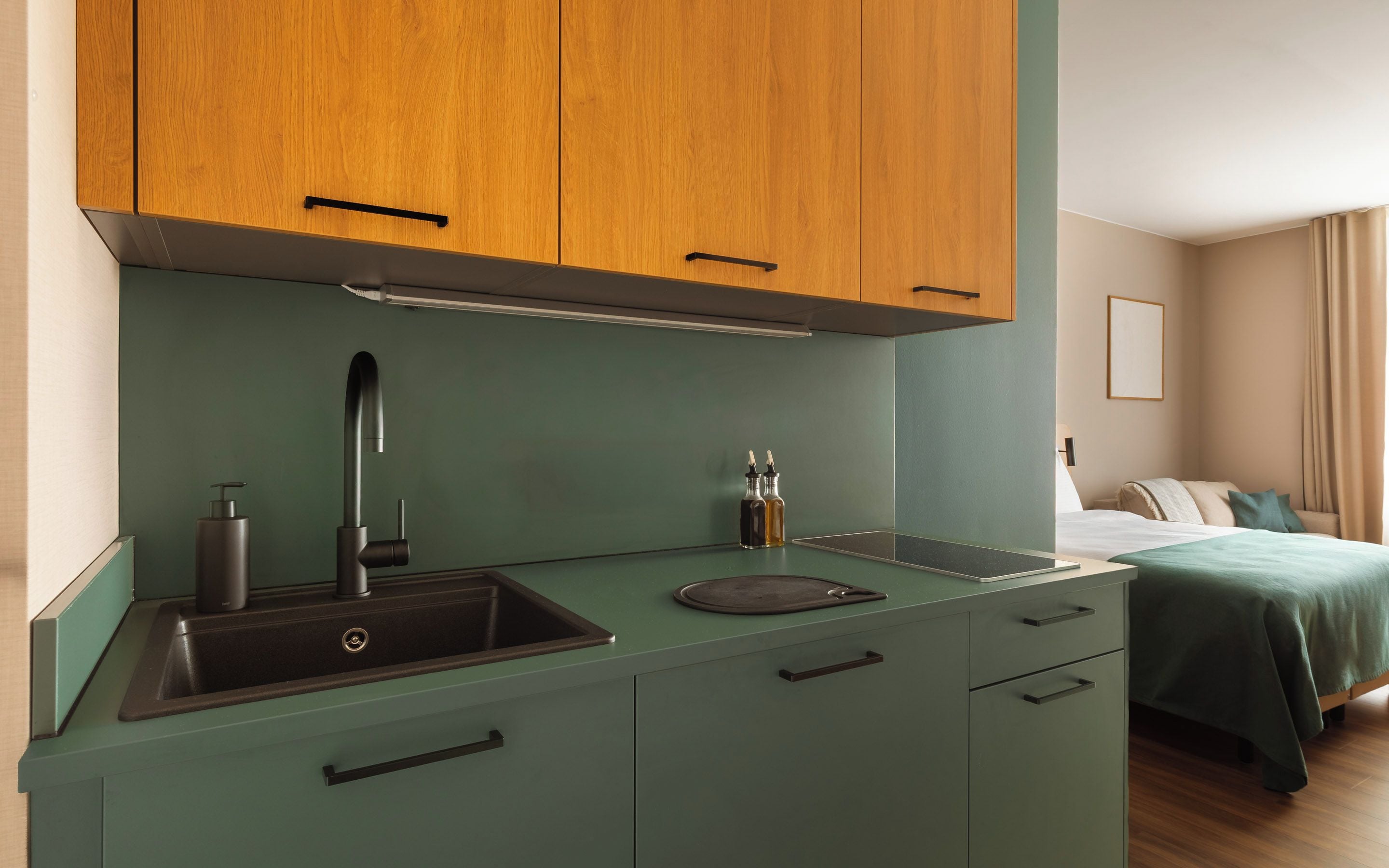 Una kitchenette compacta con un lavamanos, una grifería y una encimera. Hay armarios sobre la encimera y una cama con colcha verde al fondo.