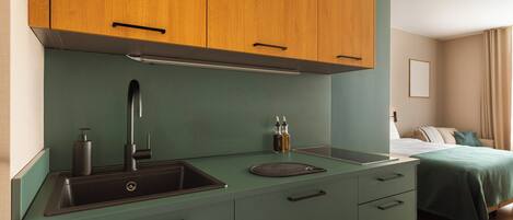Una kitchenette compacta con un lavamanos, una grifería y una encimera. Hay armarios sobre la encimera y una cama con colcha verde al fondo.