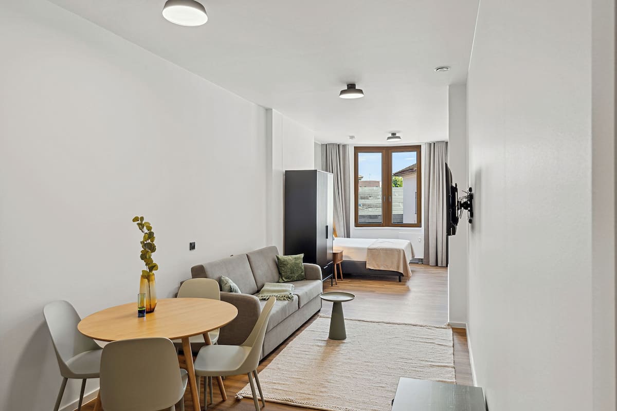 Una sala de estar moderna con sofá, mesa de comedor y sillas. Hay un televisor montado en la pared y una ventana con cortinas.
