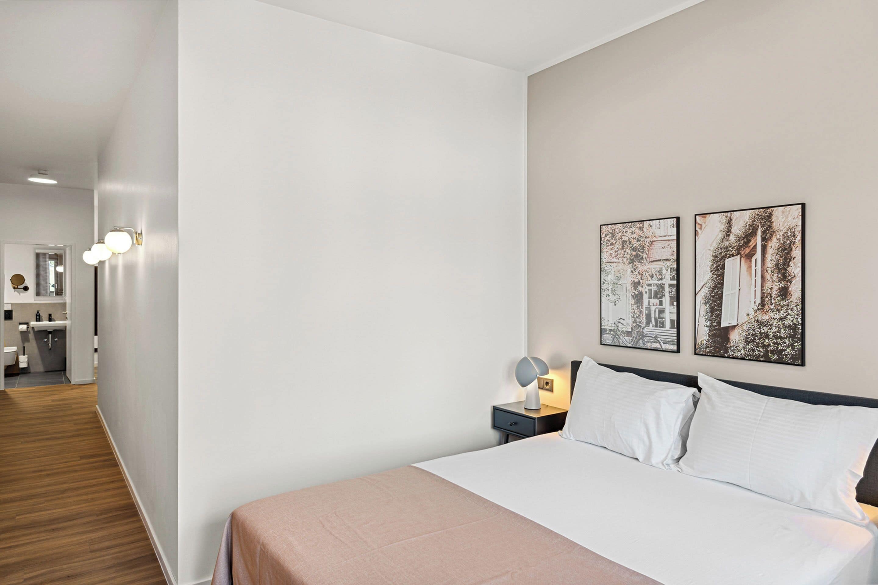 A modern hotel room with a neatly made bed, a bedside table with a lamp, and two framed pictures on the wall.