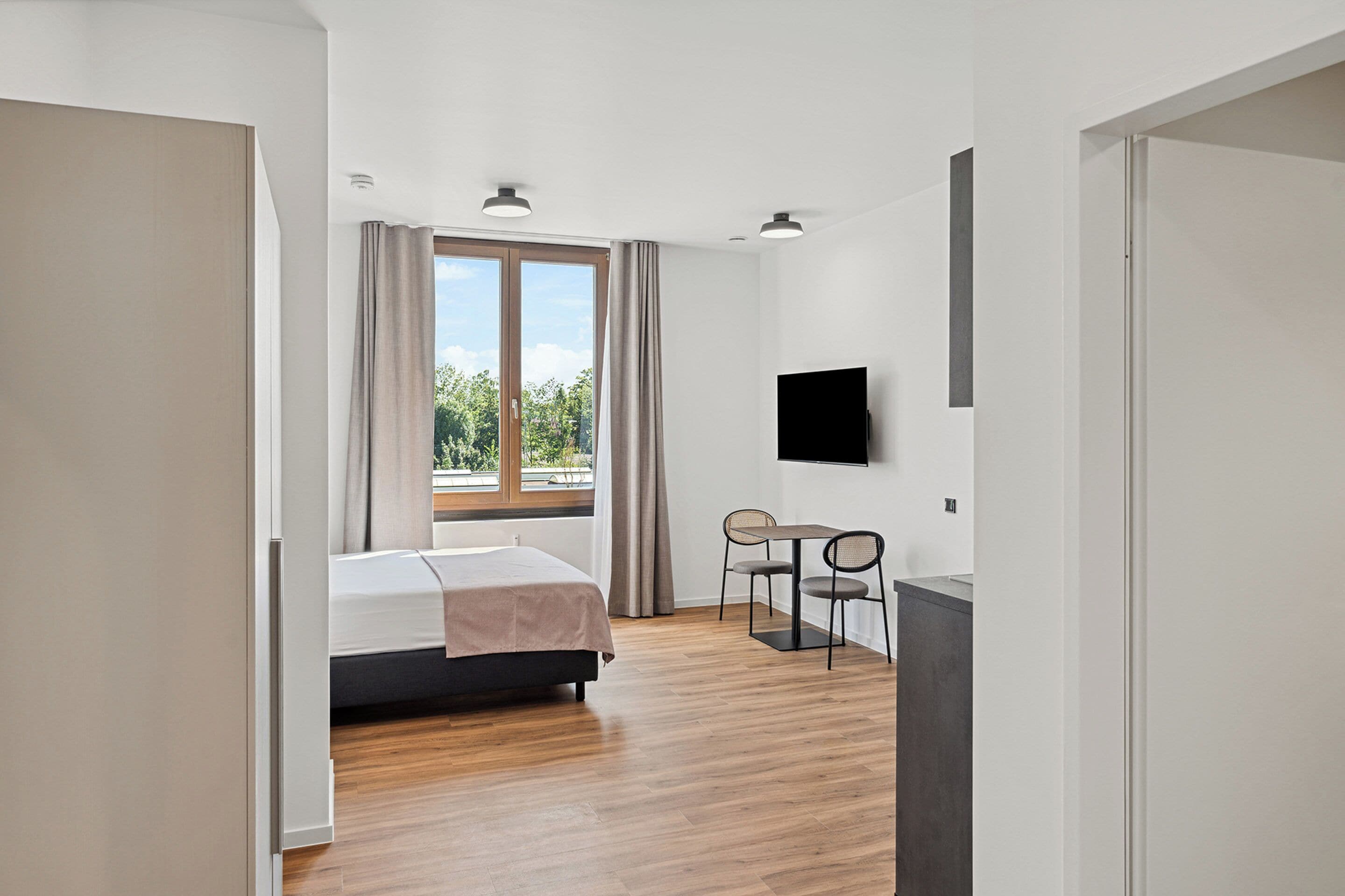 Habitación de hotel moderna con cama, escritorio, silla, televisión y un amplio ventanal con cortinas.