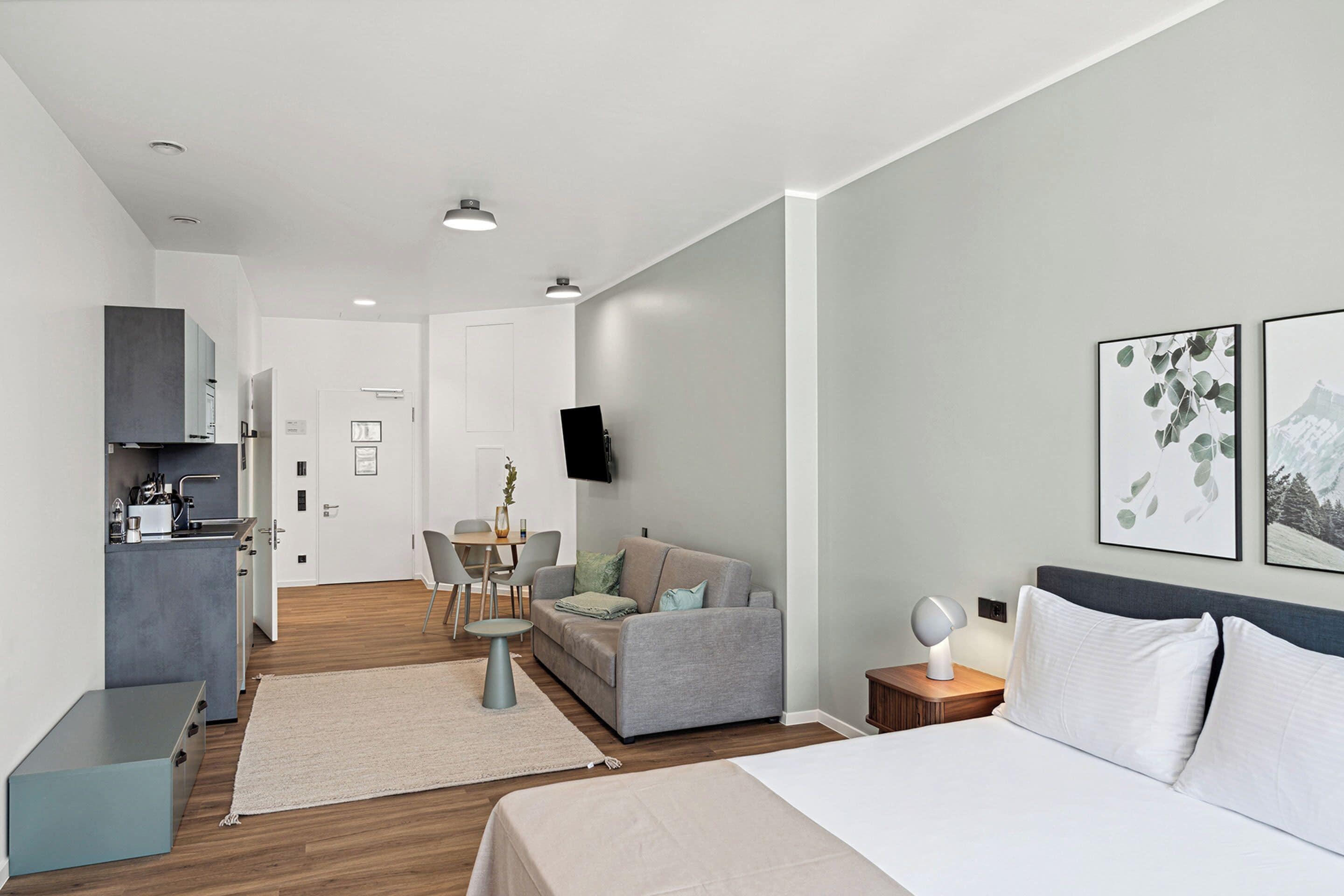 Quarto de hotel moderno com cama, sofá, área de refeições e cozinha compacta.
