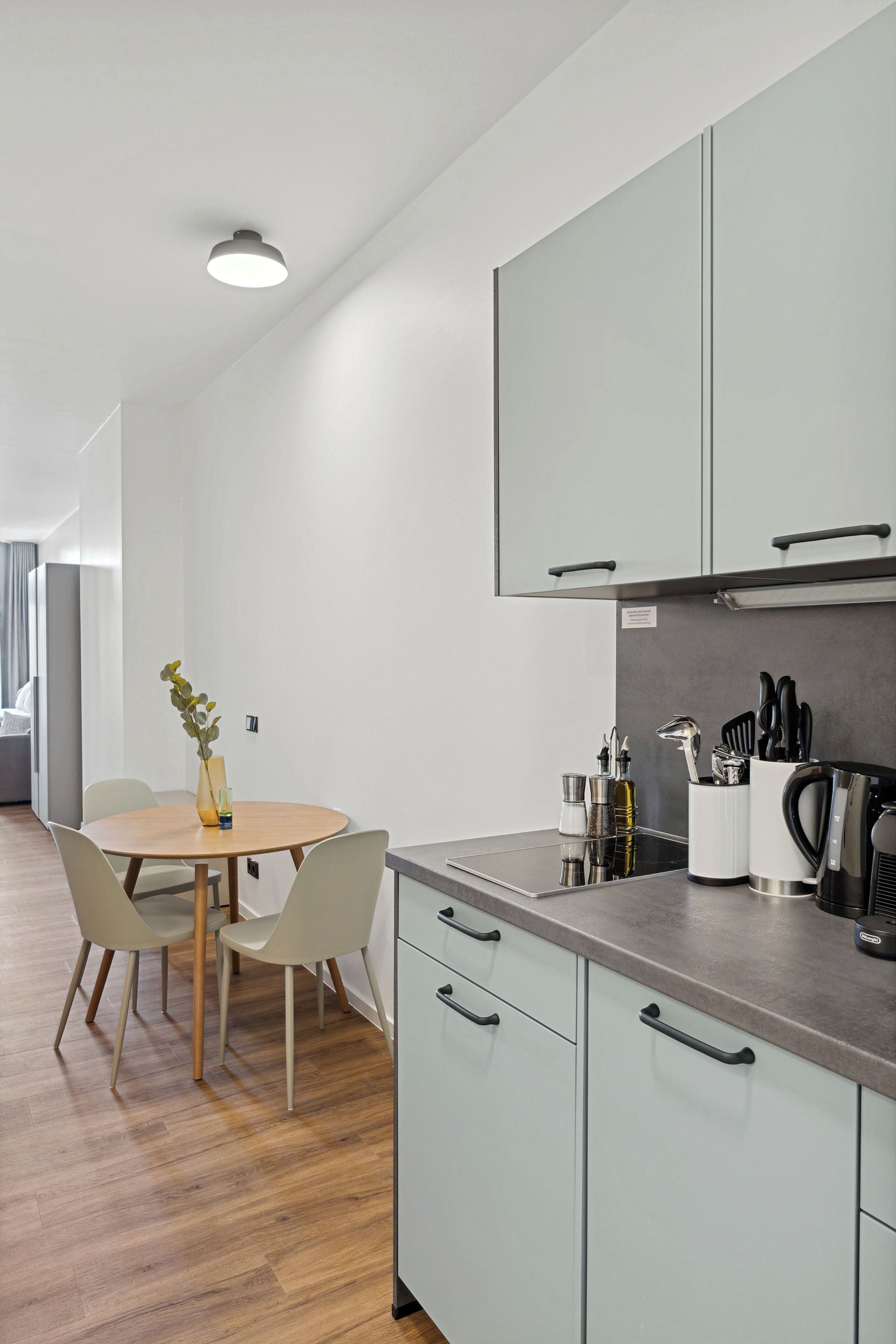 Una cocina moderna con una mesa redonda para comer, sillas de color claro y un jarrón con una planta. Se observan armarios con pomos, una cafetera y un soporte para cuchillos en la encimera.