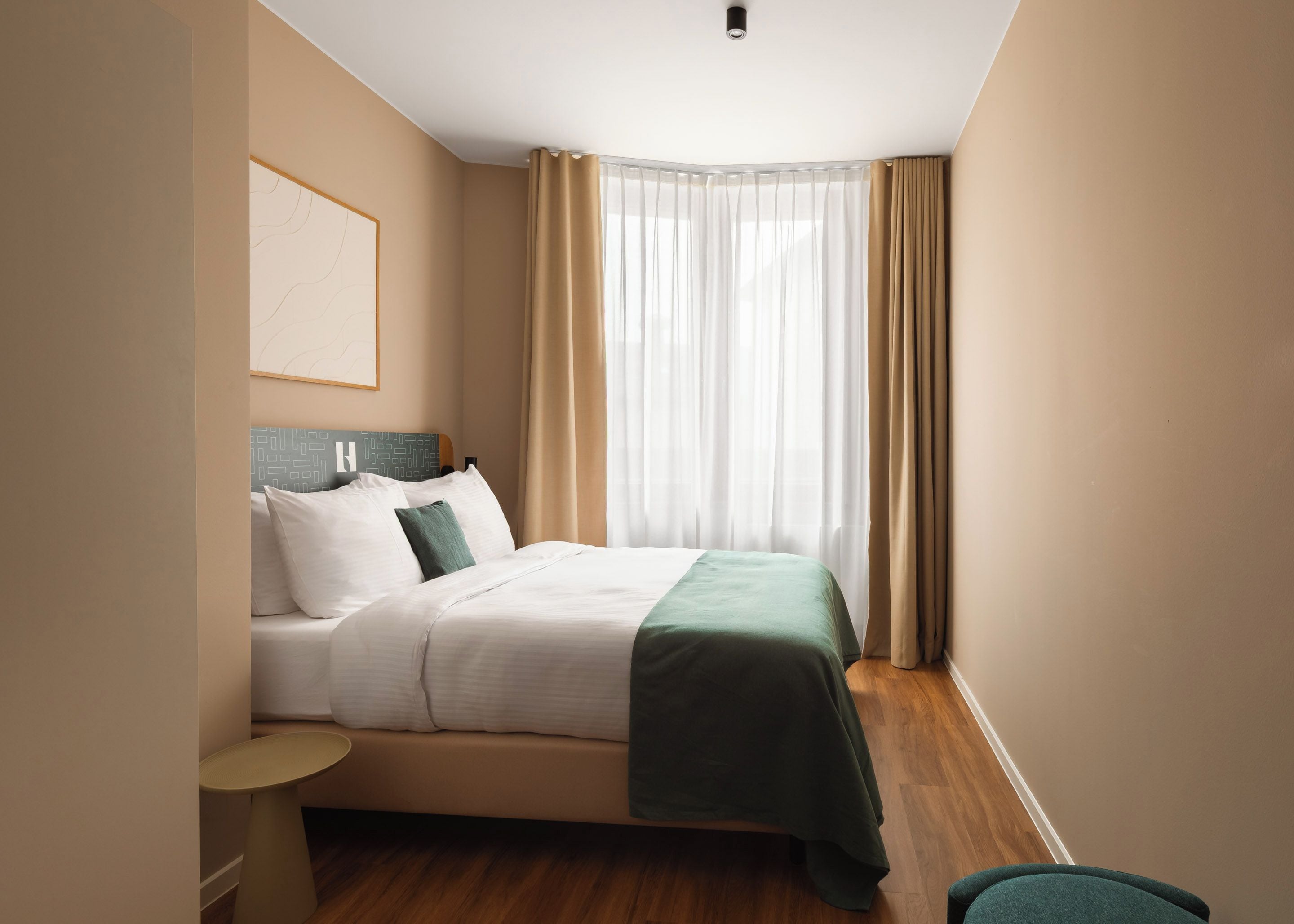 A hotel room with a bed, a bedside table, a nightstand, a window with curtains, and a wall-mounted light fixture.