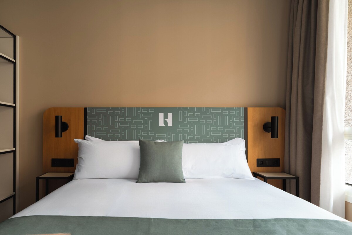 A hotel room with a bed, two bedside tables, a headboard with a geometric pattern, and a window with curtains.
