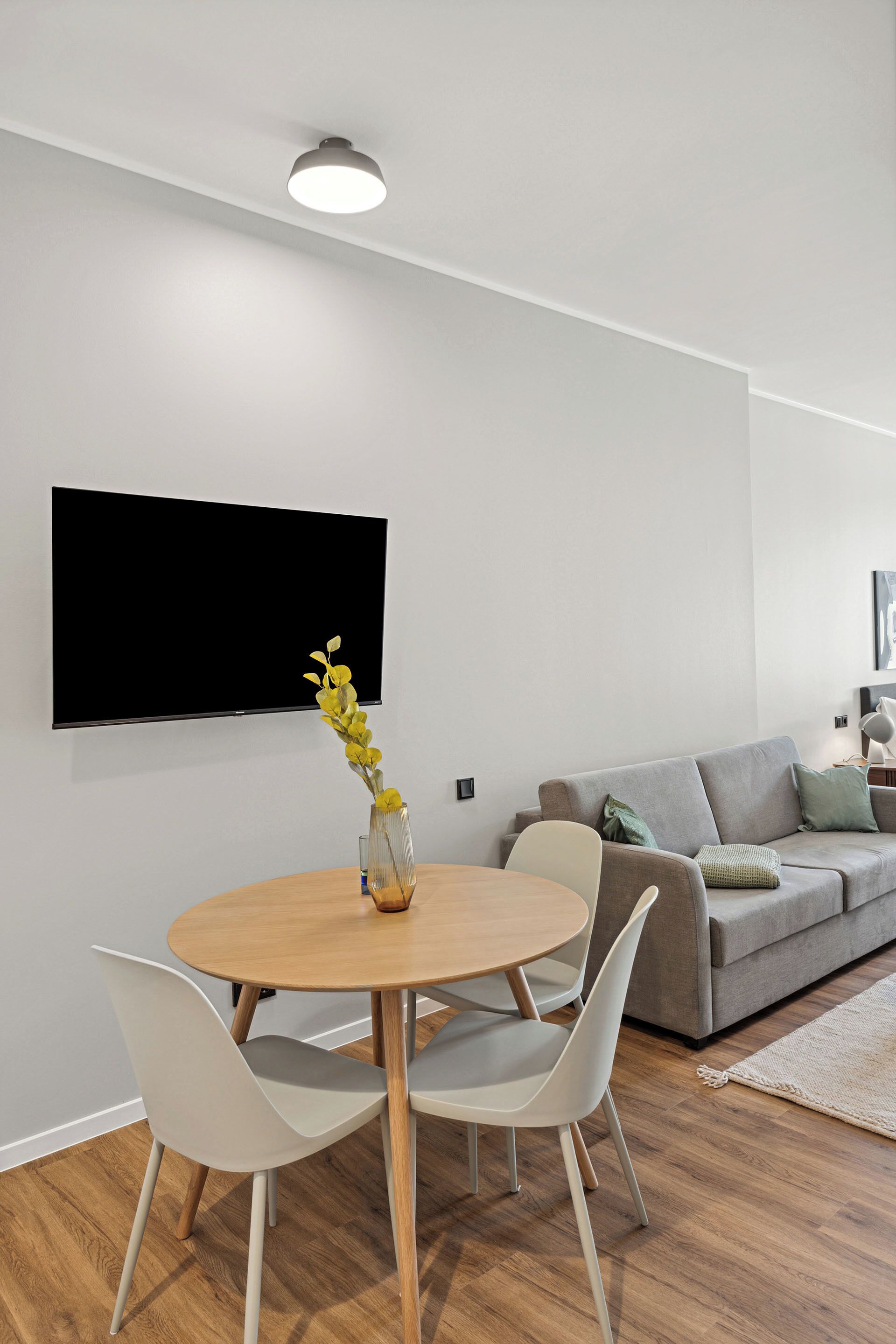 A modern living room with a round wooden dining table, two white chairs, a grey sofa, and a flat-screen TV mounted on the wall.