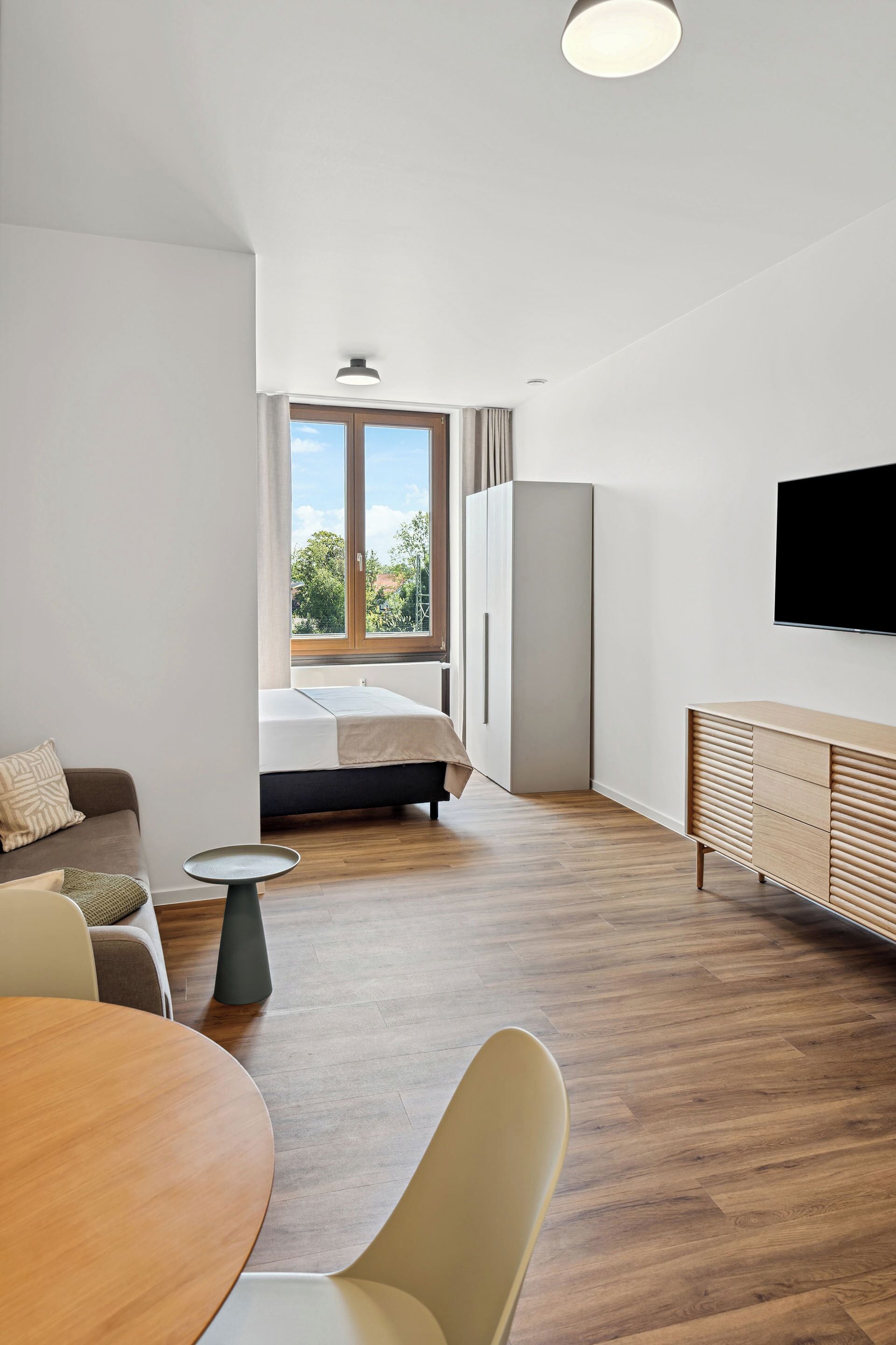 Ein modernes Hotelzimmer mit Holzboden, einem Bett mit weißem Kopfteil, einer Couch, einem Esstisch und einem an der Wand befestigten Fernseher.