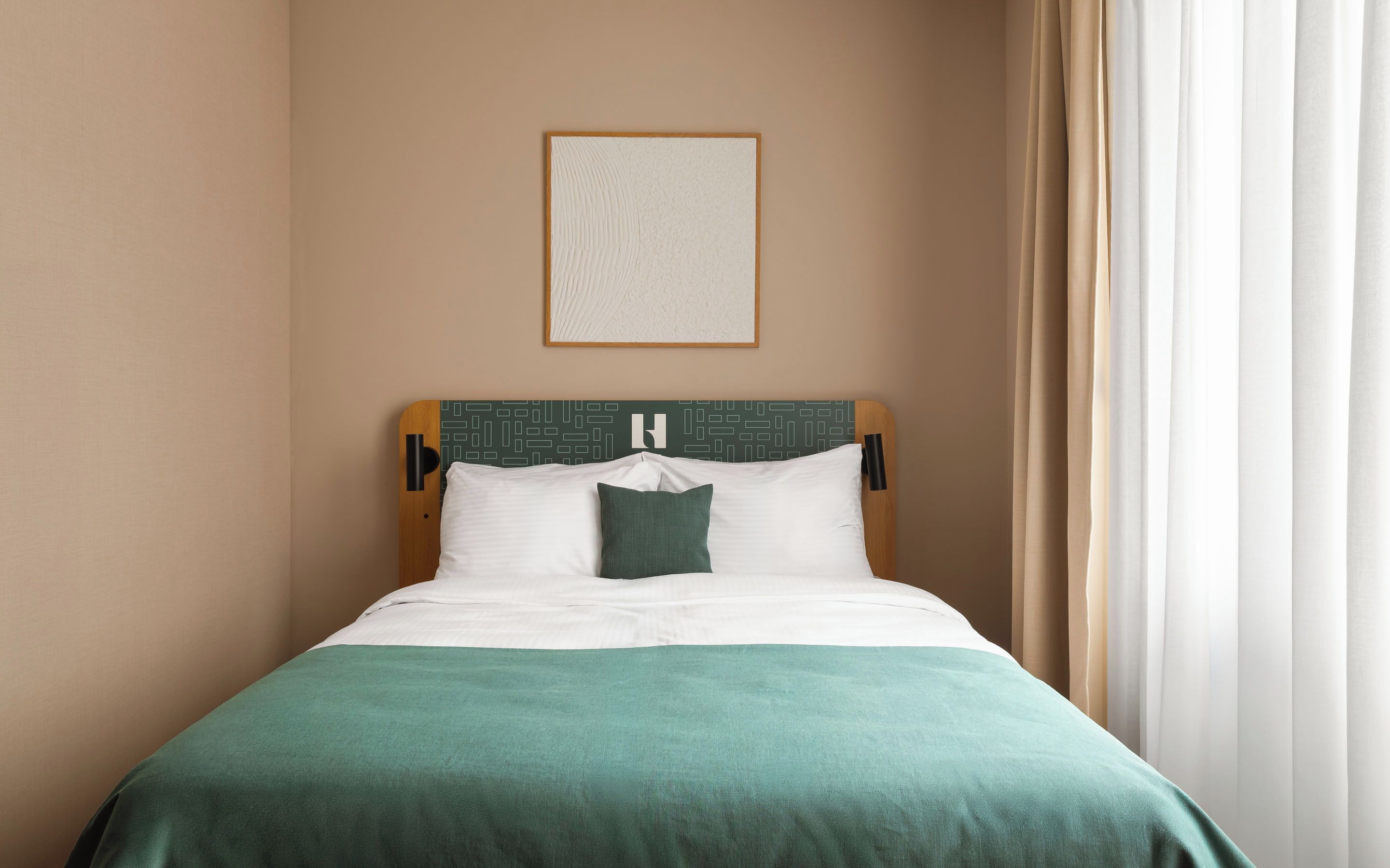 Cama bem arrumada com colcha verde e almofadas brancas, cabeceira com padrão geométrico e quadro na parede.