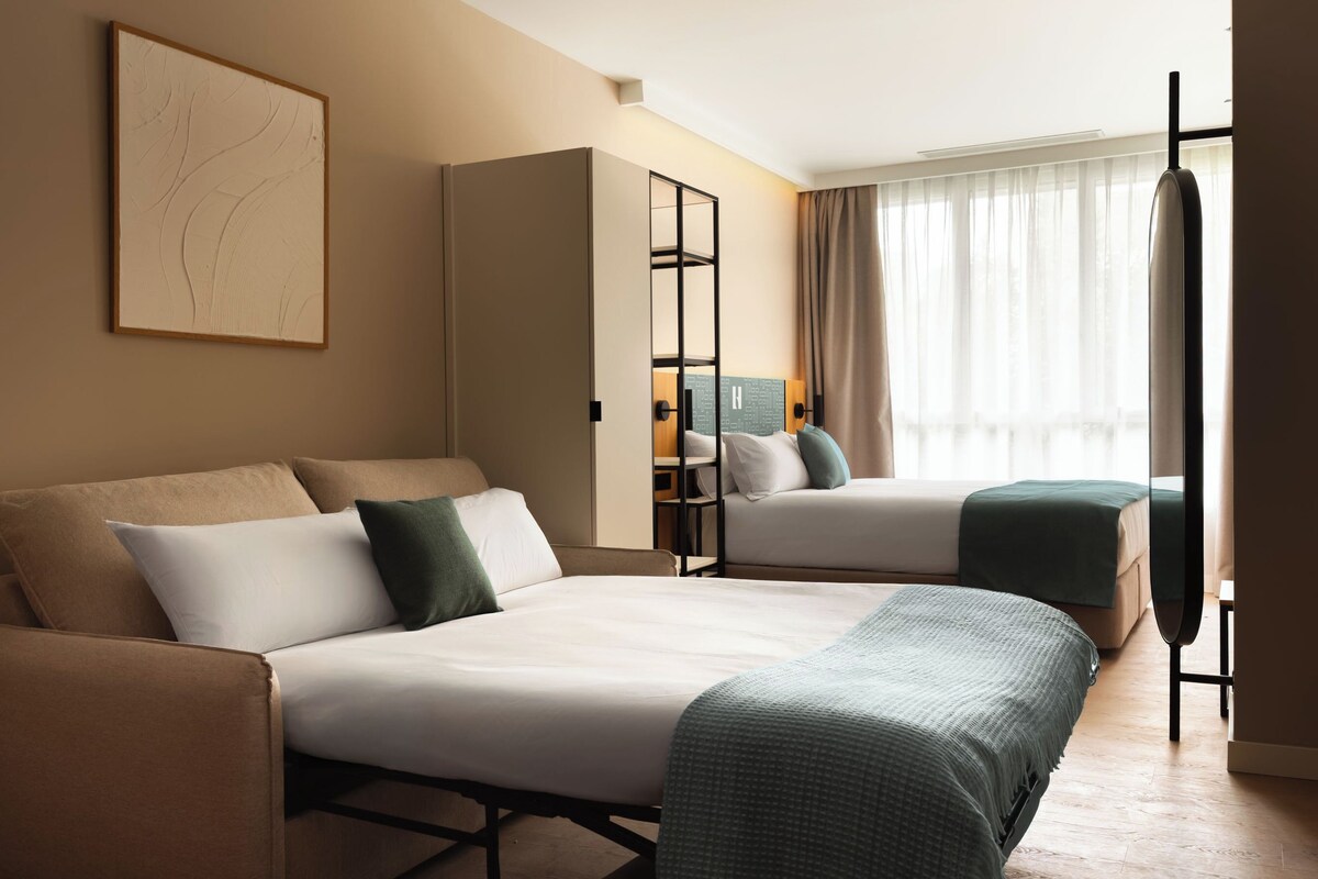 A hotel room with two beds, a sofa, a wardrobe, and a large window with curtains.