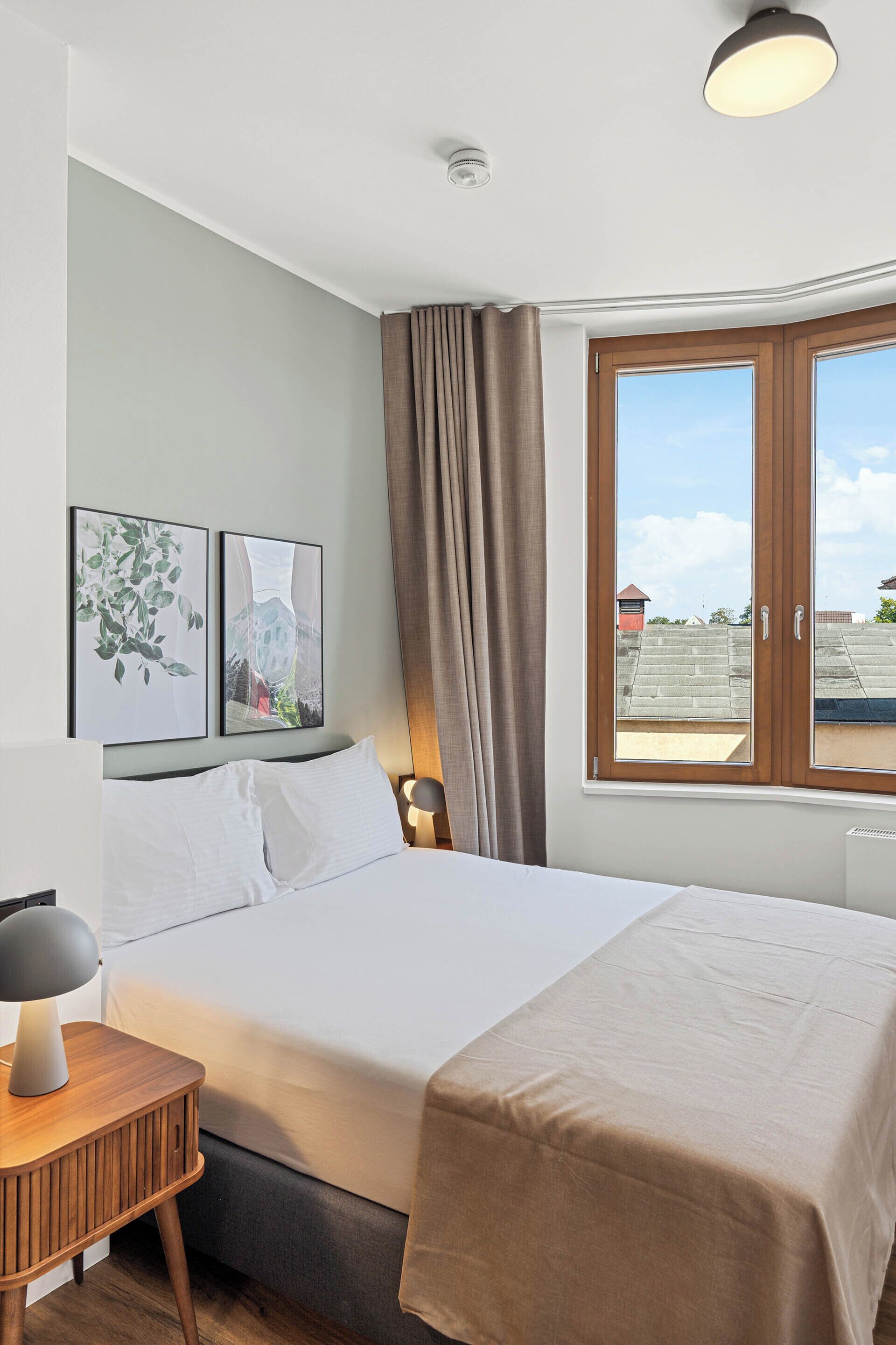 Un lit bien fait, recouvert d’une couverture blanche et beige, une table de chevet avec une lampe, et une fenêtre avec des rideaux.