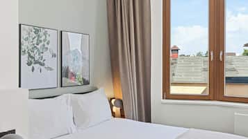 Un lit bien fait, recouvert d’une couverture blanche et beige, une table de chevet avec une lampe, et une fenêtre avec des rideaux.