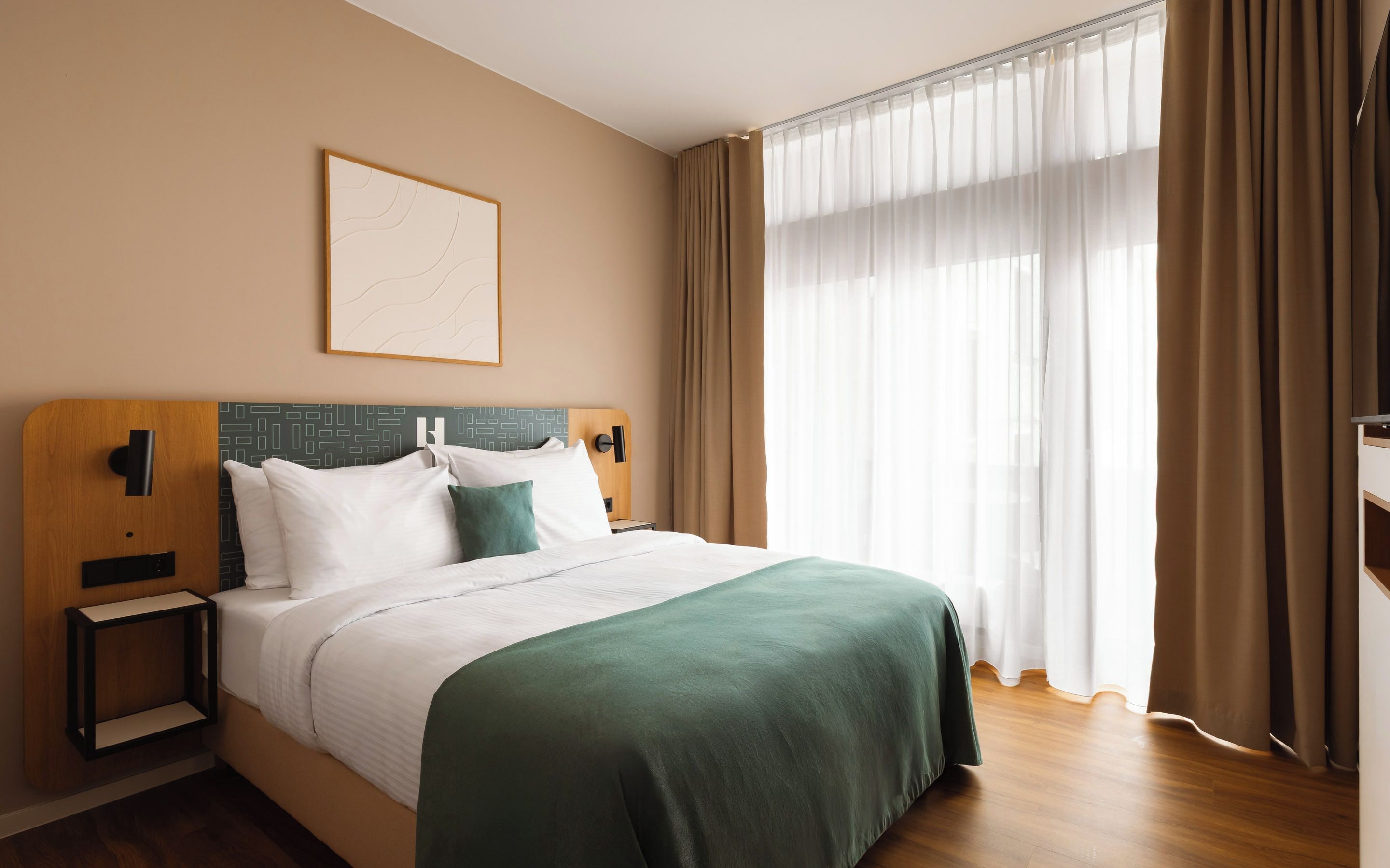 A hotel room with a bed, bedside tables, a nightstand, a wall-mounted artwork, and sheer curtains.