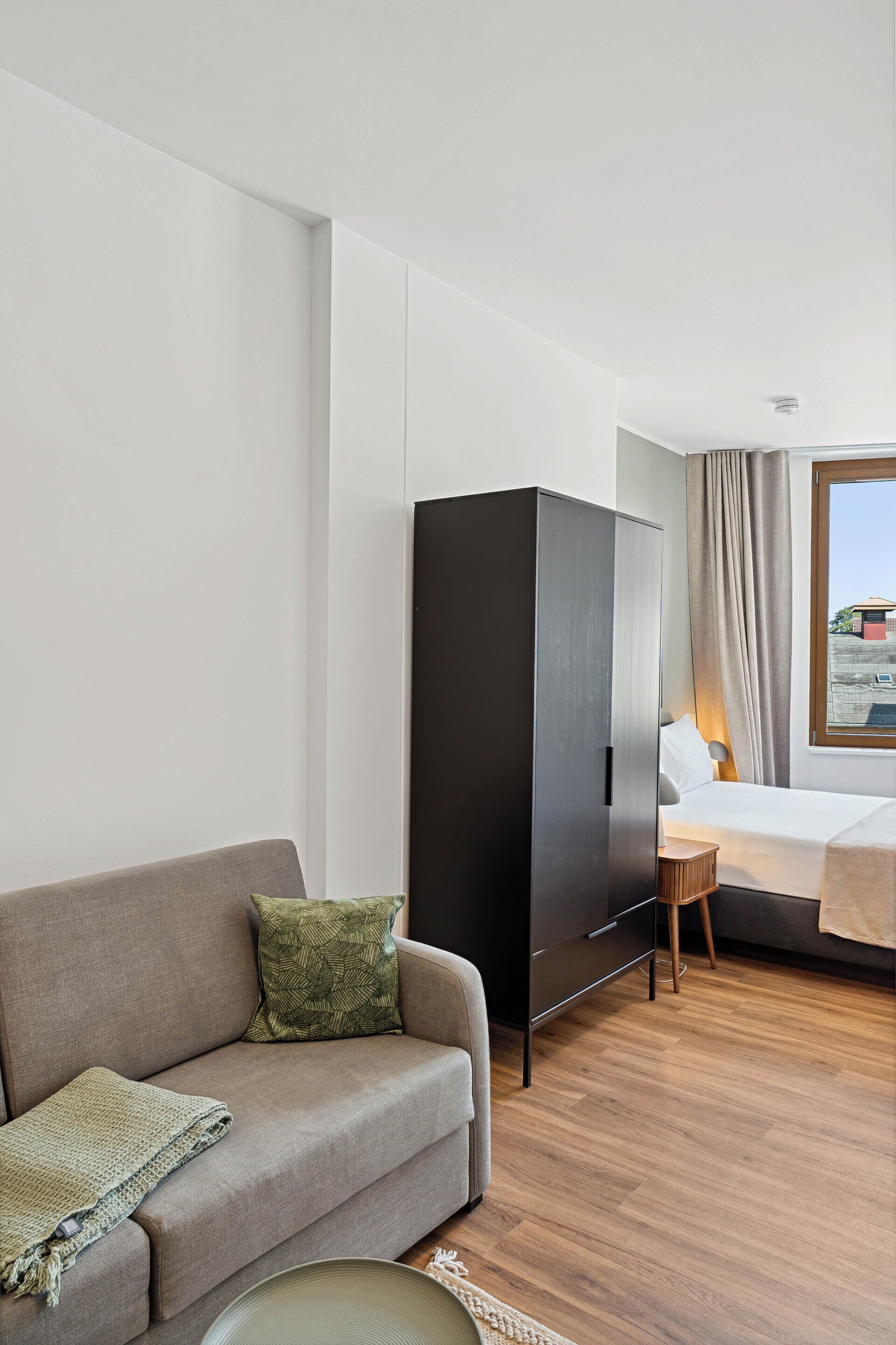 A modern hotel room with a grey sofa, a wooden floor, a wardrobe, a bed with a white headboard, and a window with curtains.