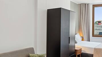 A modern hotel room with a grey sofa, a wooden floor, a wardrobe, a bed with a white headboard, and a window with curtains.