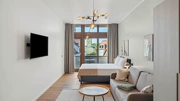 A modern hotel room with a large bed, a sofa, a flat-screen TV, and a balcony with a view of buildings.