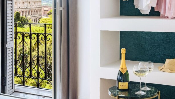 A room with a balcony, a wine bottle, and a wine glass on a small table. - Elysium Suites Colosseo (Rome)