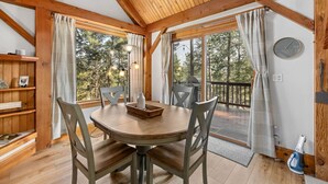 Un coin repas en bois avec une table ronde et des chaises, une horloge au mur et une vue sur les arbres à travers de grandes fenêtres.