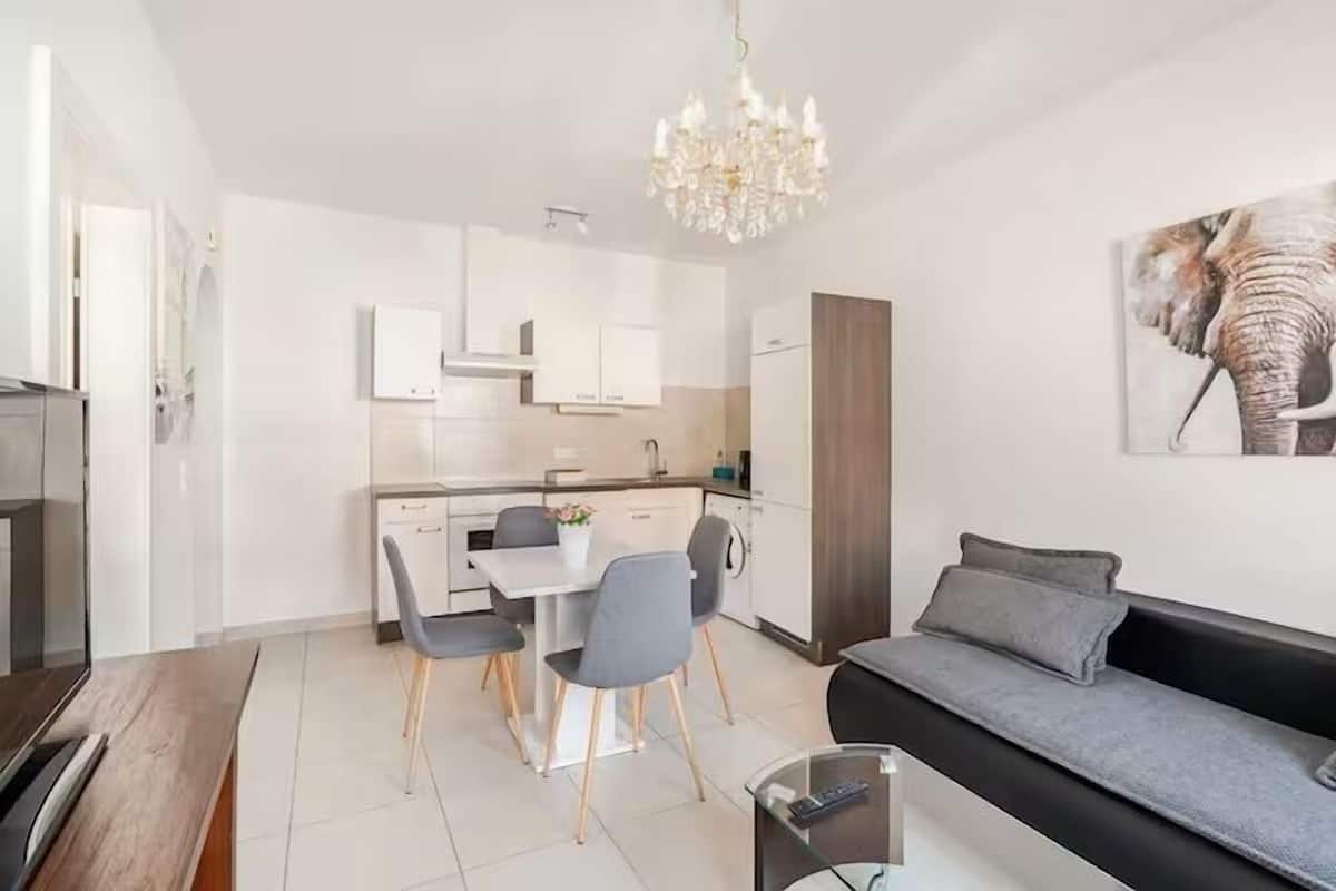 A modern living room with a sofa, a dining table with chairs, a kitchen area with cabinets, and a chandelier.