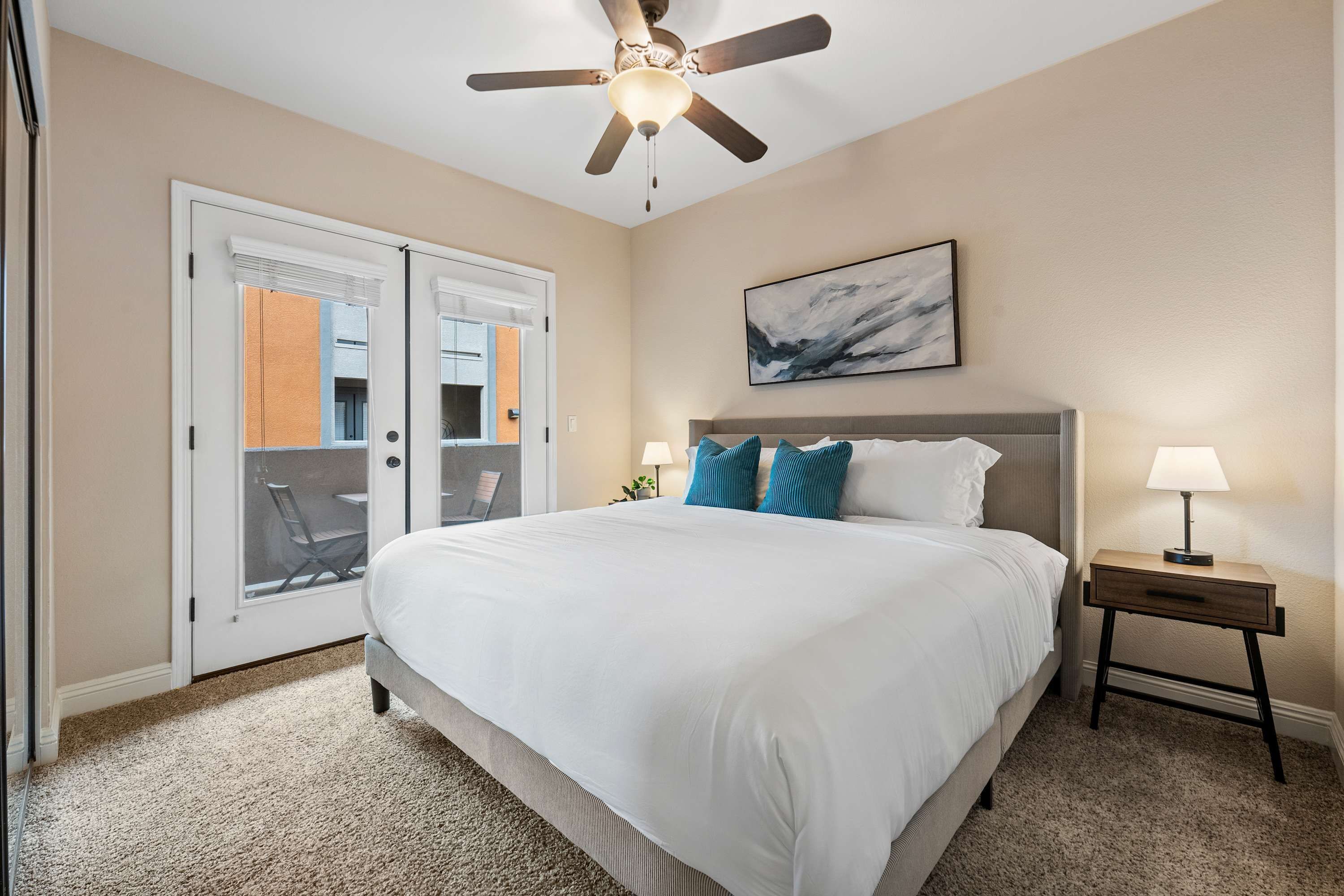A bedroom with a bed, a ceiling fan, a nightstand with a lamp, and a door leading to a balcony.