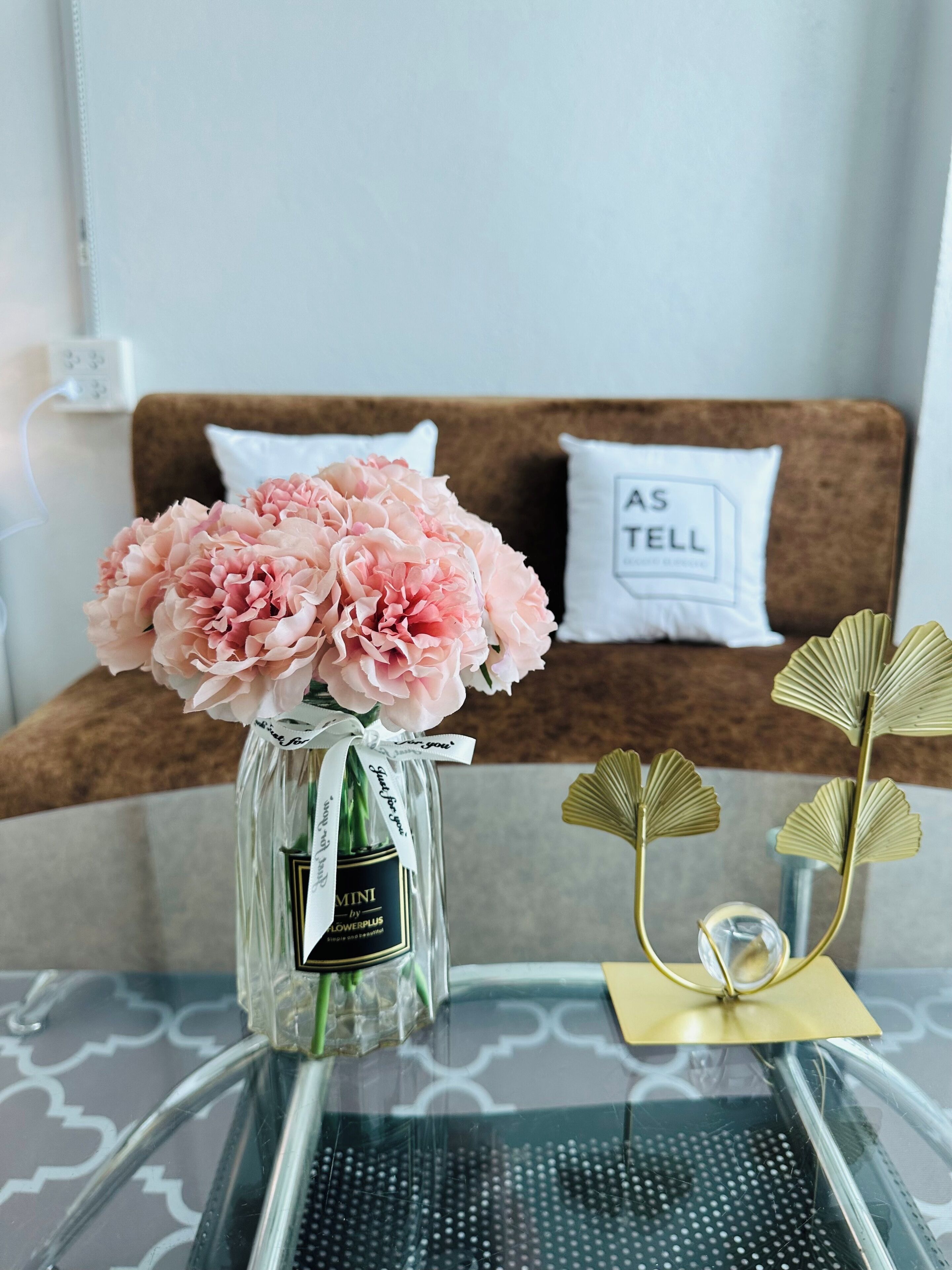 Ein Glastisch mit einer Vase voller pinkfarbener Blumen, einem dekorativen Metallobjekt und einer Couch mit Kissen in einem Raum.