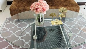 A modern living room with a brown sofa, two white pillows with "AS TELL" text, a glass coffee table with a vase of flowers, and a decorative gold leaf piece. - Service apartment lampang (Lampang)
