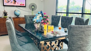 A dining area with a glass-top table, grey upholstered chairs, a floral centerpiece, and a TV displaying an advertisement. - Service apartment lampang (Lampang)