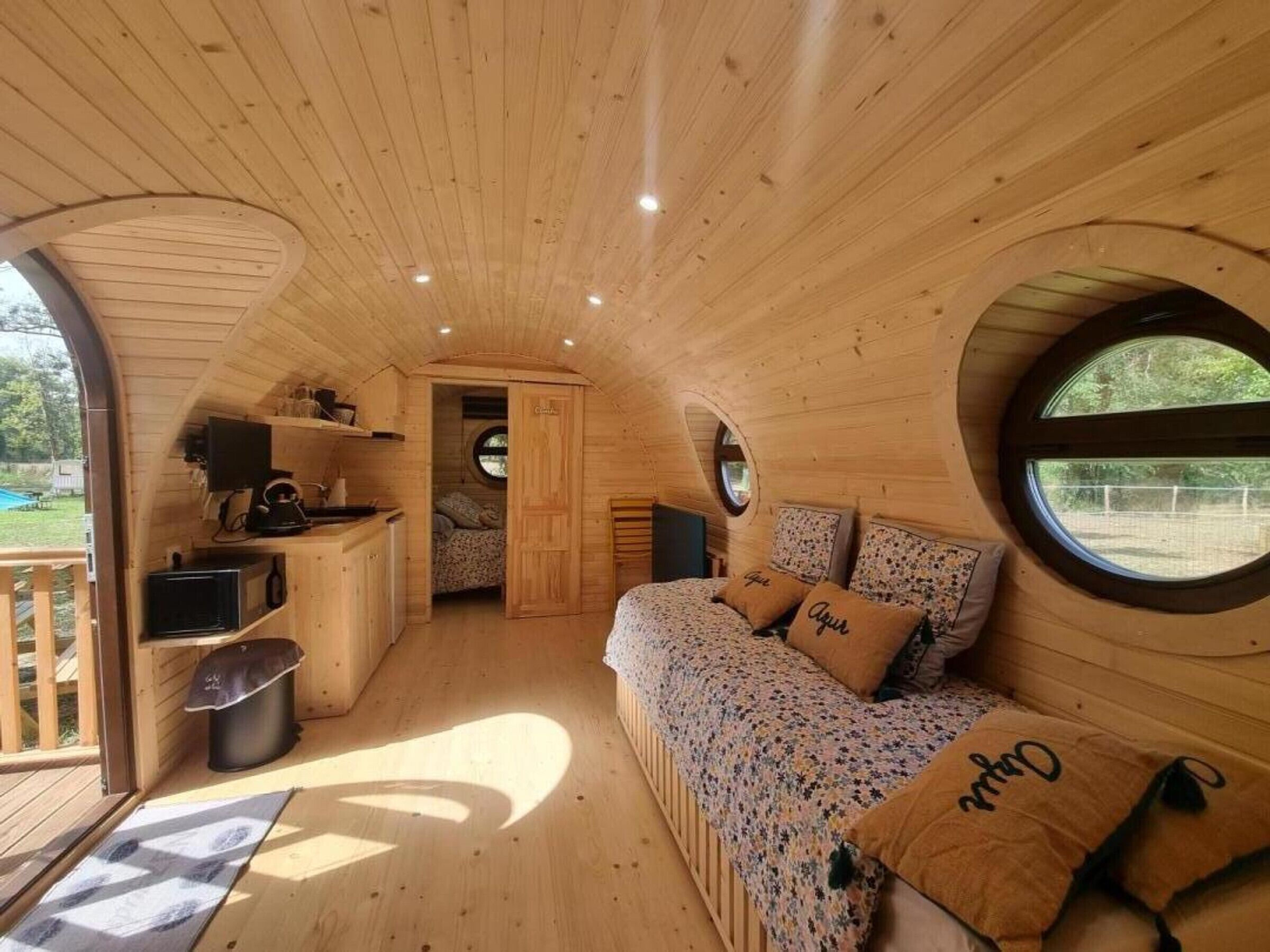 A cozy cabin interior with a wooden ceiling, a sofa with floral bedding, and a small kitchen area.
