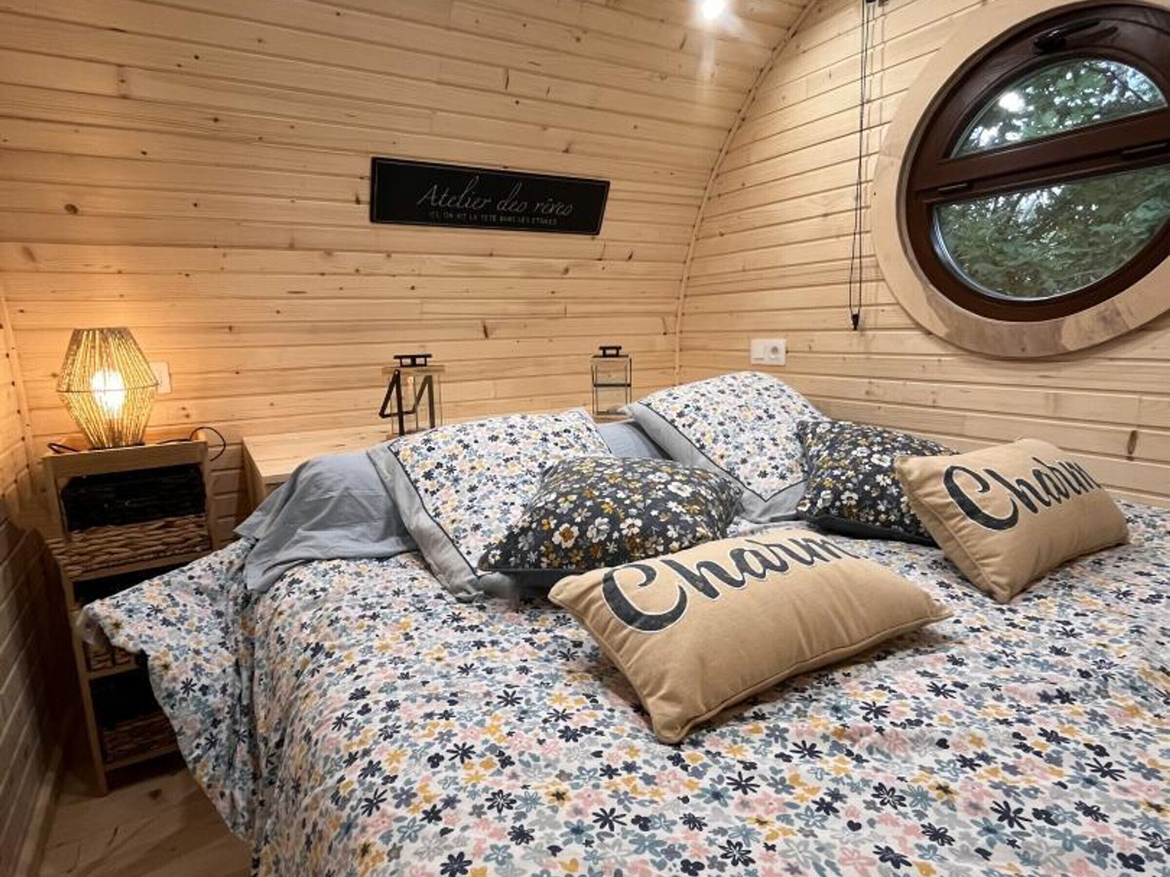Un dormitori acollidor amb interior de fusta, una finestra rodona i un llit amb roba de llit i almoinos amb motius florals.