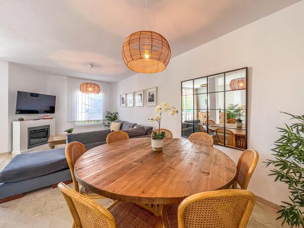 Un salon moderne avec une table à manger en bois, des chaises en osier, un canapé et un téléviseur fixé au mur.