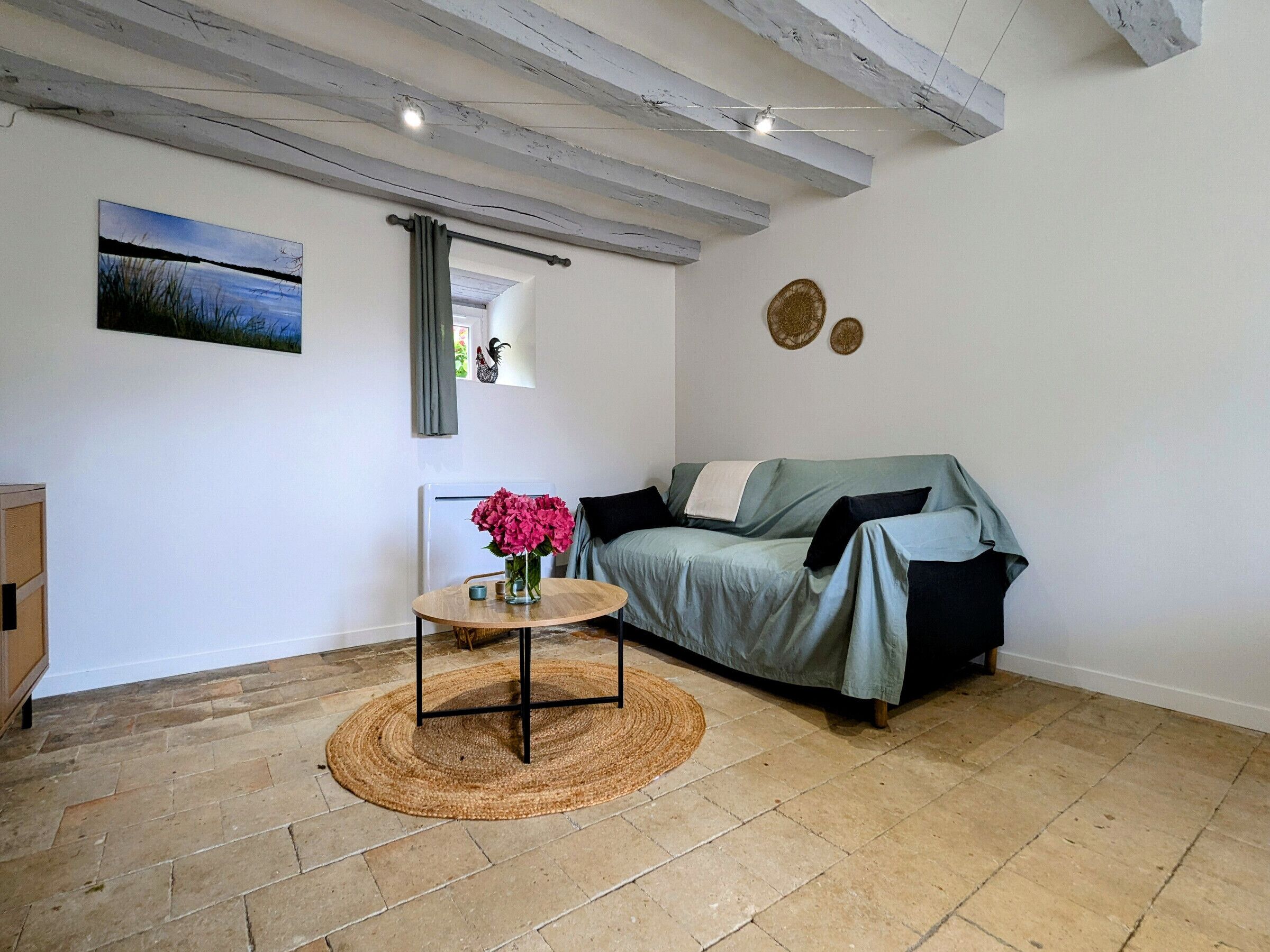 A cozy living room with a stone tile floor, a green sofa, a small round table with a vase of flowers, and a rustic ceiling with exposed beams.