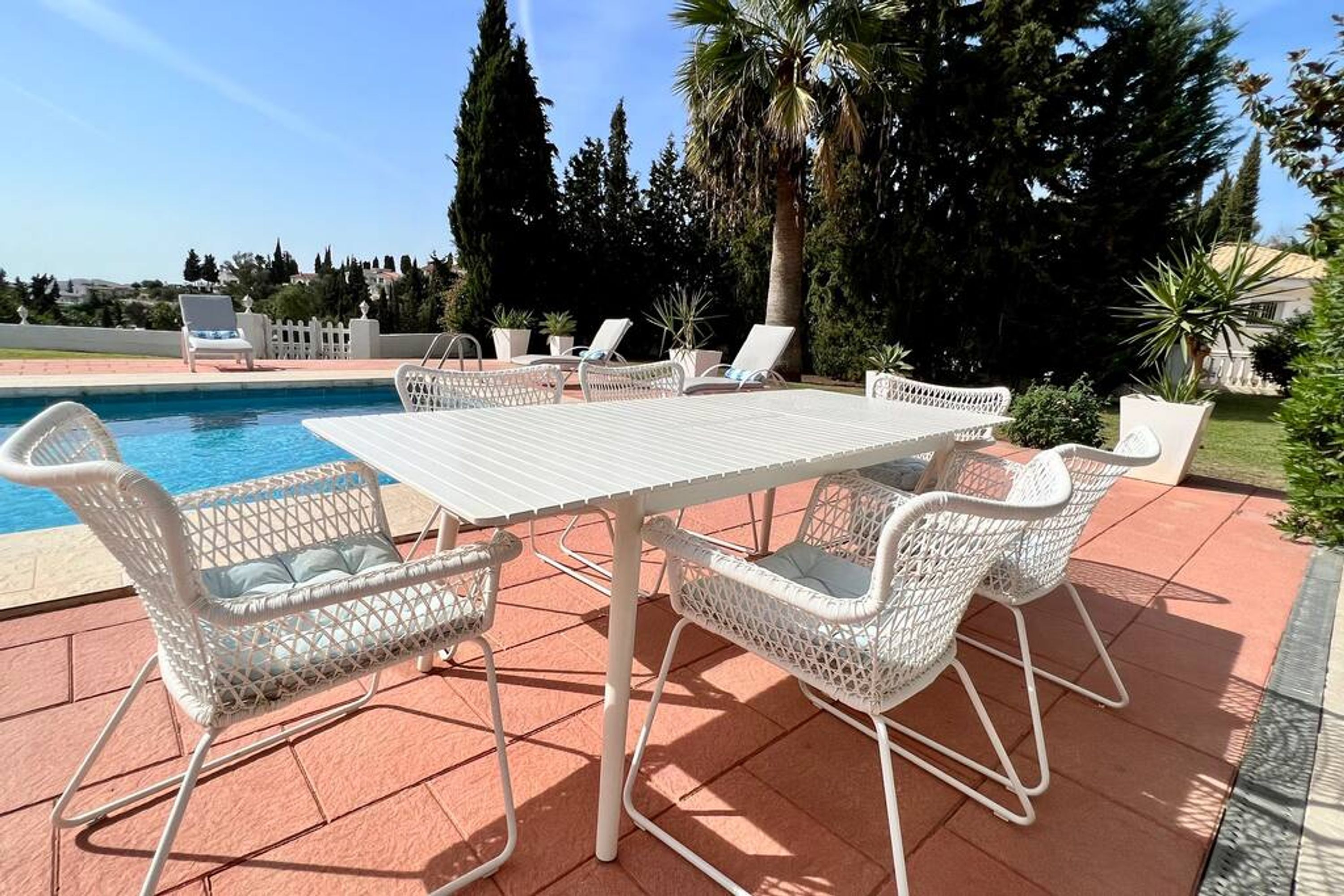 A poolside area with a white table and wicker chairs, surrounded by a tiled patio and palm trees.