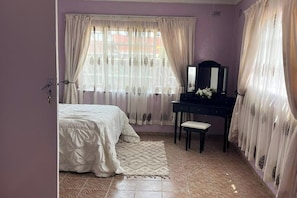 A bedroom with a bed, a dresser, a mirror, and a window with curtains. - 4 Bed Gated House-sleeps 8-private Parking (Harare)