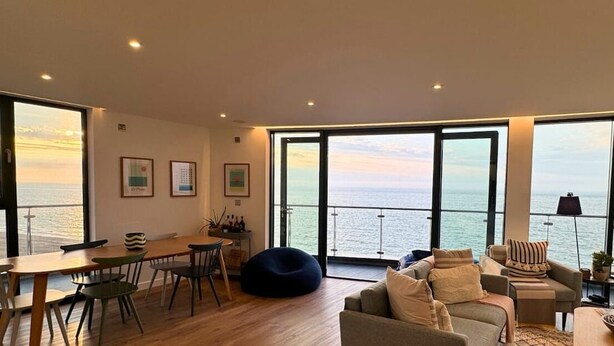 Amplia sala de estar con ventanas grandes que ofrecen vista al mar. Cuenta con una mesa de comedor con sillas, un sofá y un sillón de peluche.