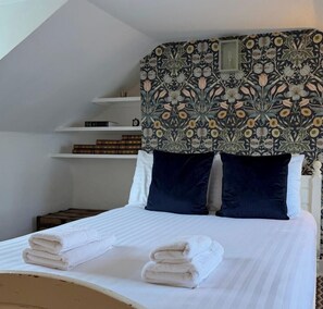 Ein ordentlich bezogenes Bett mit weißen Leinen und gefalteten Handtüchern, ein dunkles Kopfstück mit Blumenmuster und eine weiße Wand mit einem Regal, das Bücher und Kleinigkeiten beherbergt.