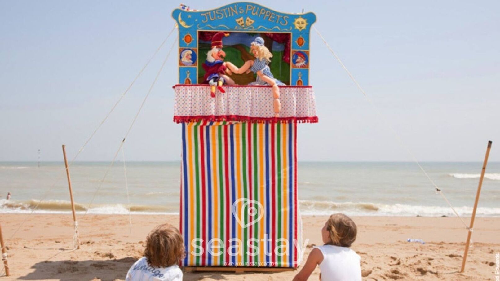 A beach scene with a striped beach umbrella and a puppet show tent.