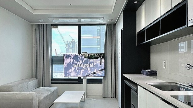 A modern hotel room with a minimalist design, featuring a sofa, a small table, a kitchen area with a sink and cabinets, and a large window offering a city view.
