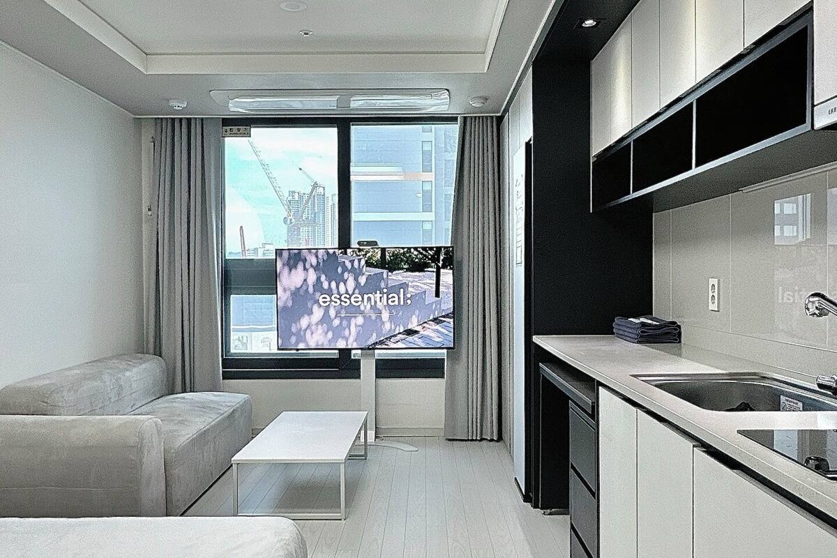 A modern hotel room with a minimalist design, featuring a sofa, a small table, a kitchen area with a sink and cabinets, and a large window offering a city view.